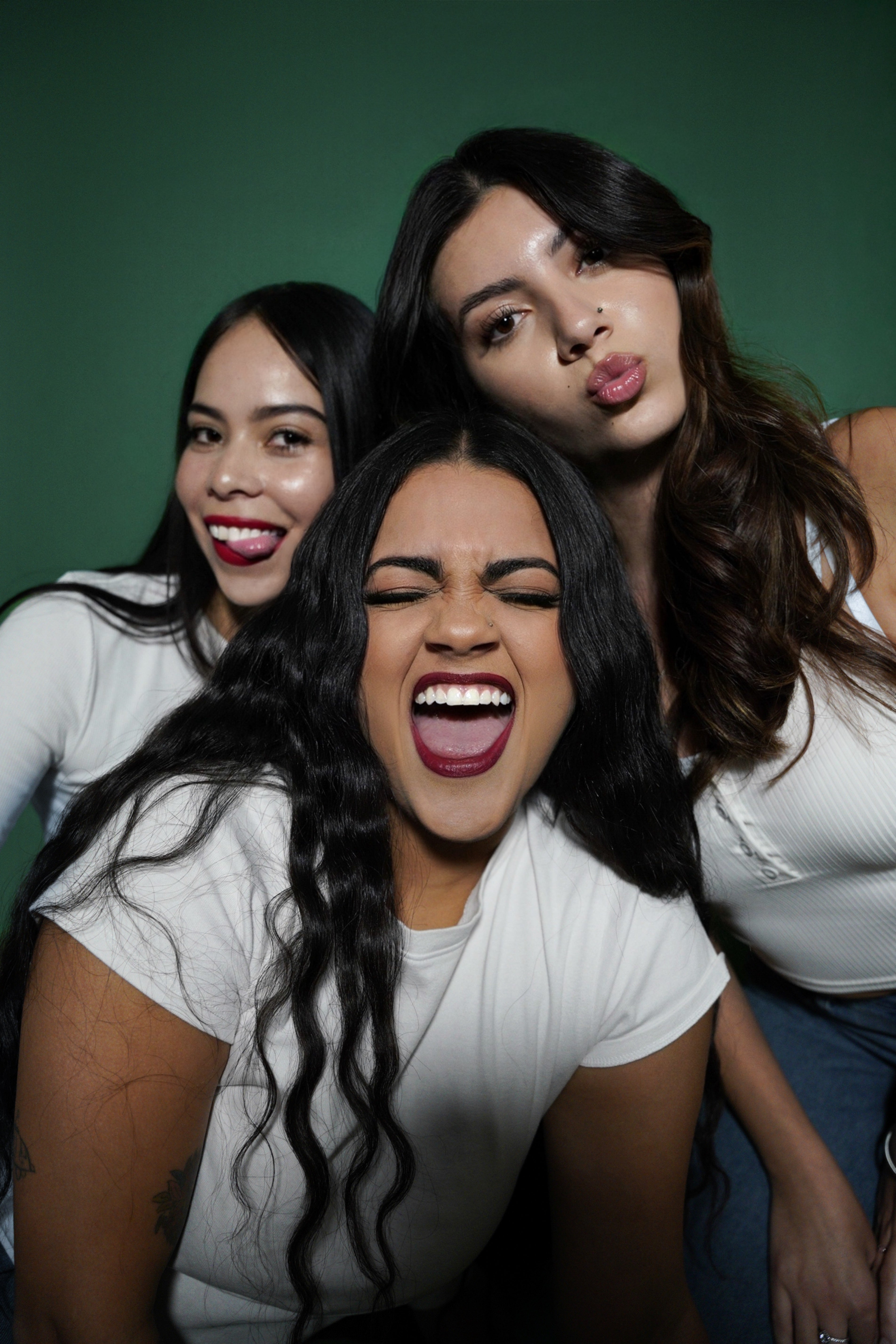 Three women posing together with a green background wearing lipstick and lip gloss from the sasso cosmetics collecion